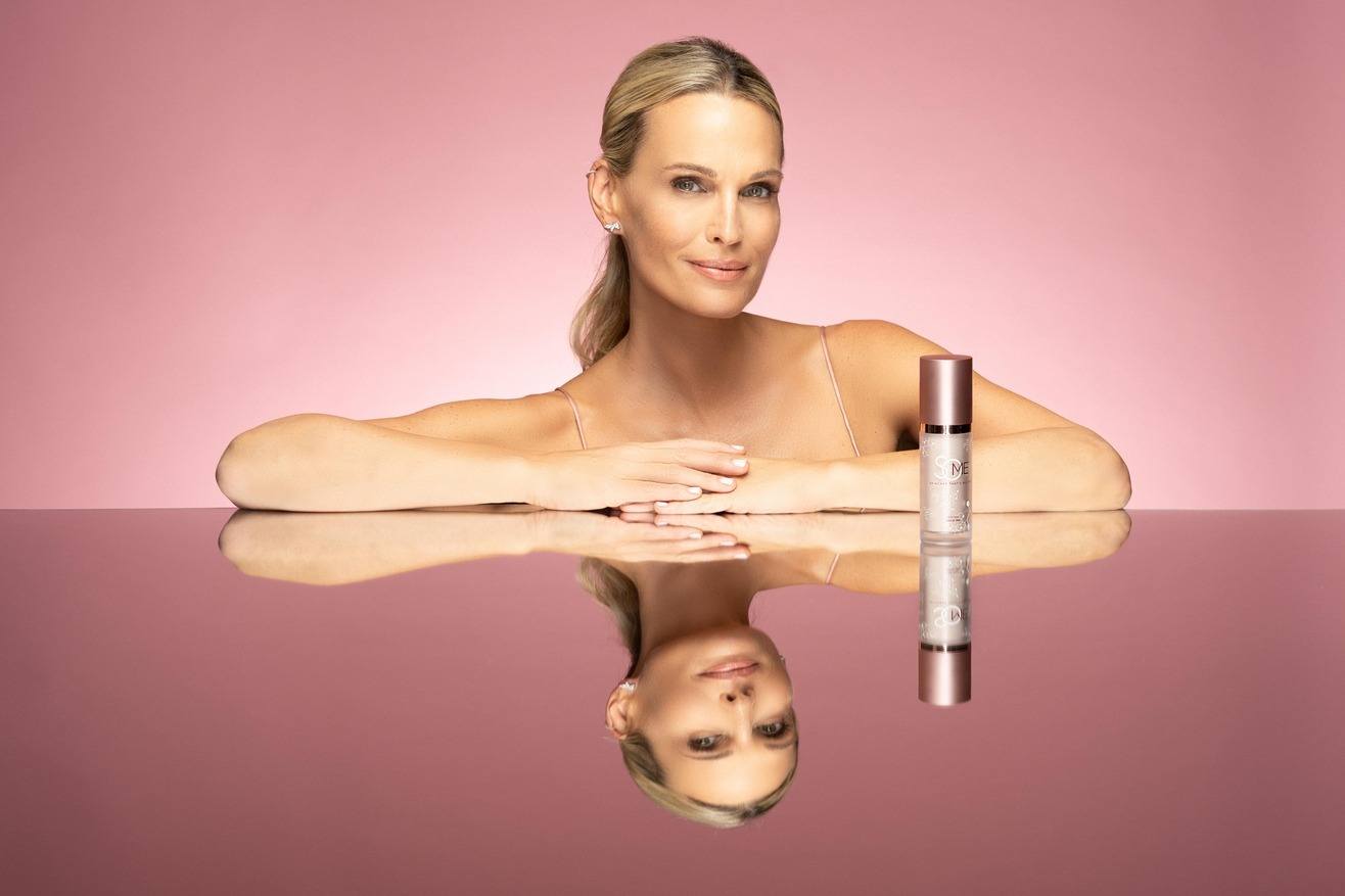 Molly Sims with flawless skin poses with a bottle of SoME Skincare against a pink background.